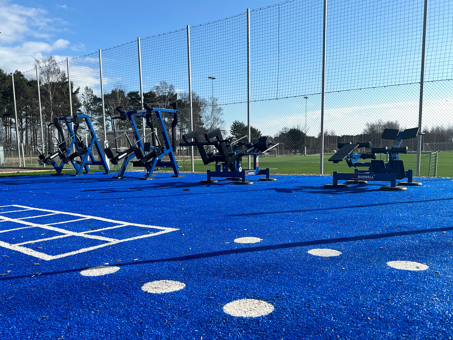 Utomhusgym med styrketräningsutrustning placerat intill en fotbollsplan på en idrottsanläggning, utformat för träning och fysisk aktivitet i offentlig miljö.