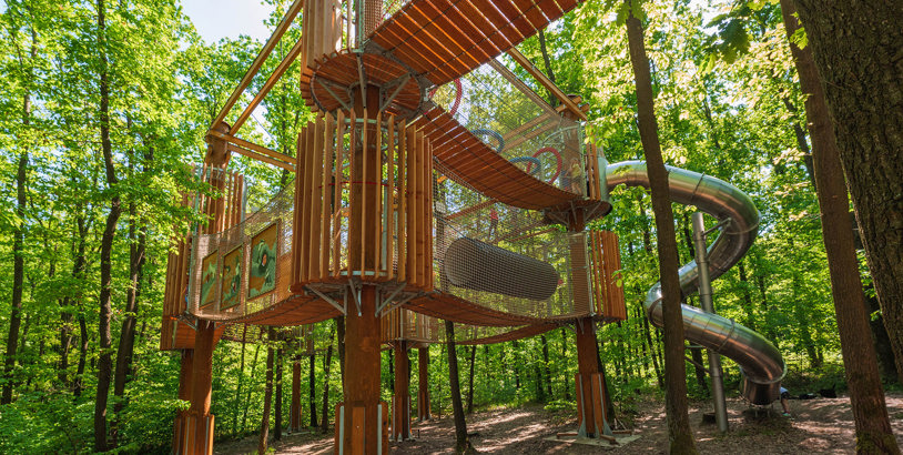 Amaze signaturlekplats i skogsmiljö med upphöjda träkonstruktioner, nätgångar och spiralrutschbana som skapar ett lekfullt landmärke bland träden.