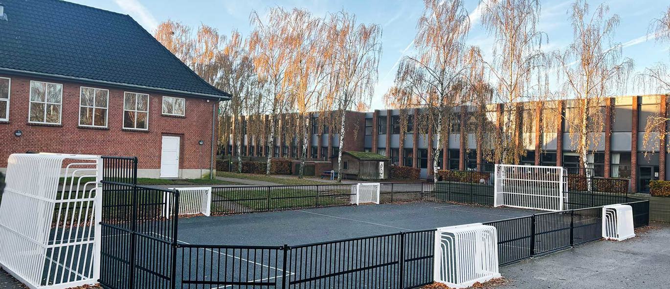 Multibane med svarte vant og hvite mål på lang- og kortsidene, plassert på asfalt med skolebygninger i bakgrunnen.