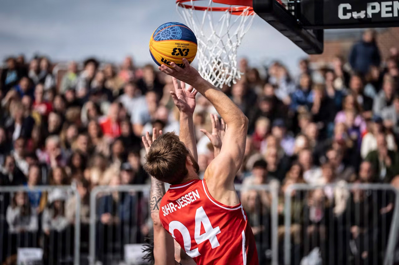 en basketballspiller spiller en gul og blå basketball opp mot et basketballnett i en rød og hvit sportstrøye med et stort publikum i bakgrunnen