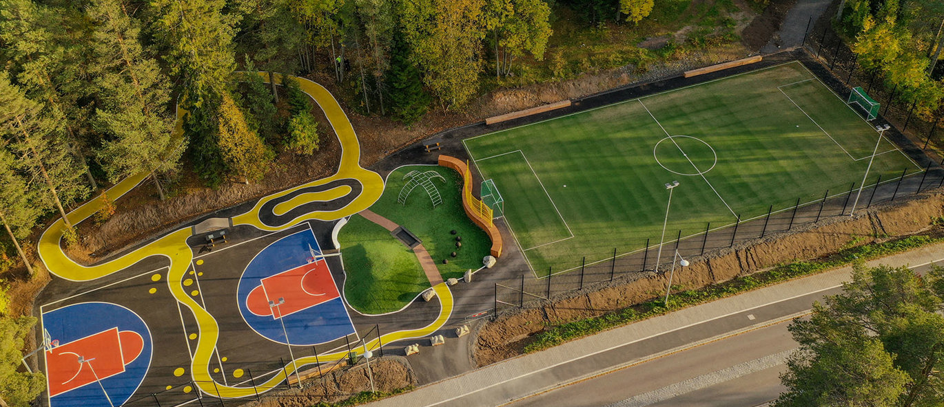 Stort idrætsområde i park omgivet af træer med multibane, basketbane, legeplads og asfaltopstregninger