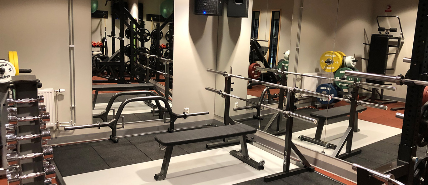 Innegym i Gällivare ishall med bänkpress, skivstänger, fria vikter och spegelvägg i kompakt träningsmiljö.