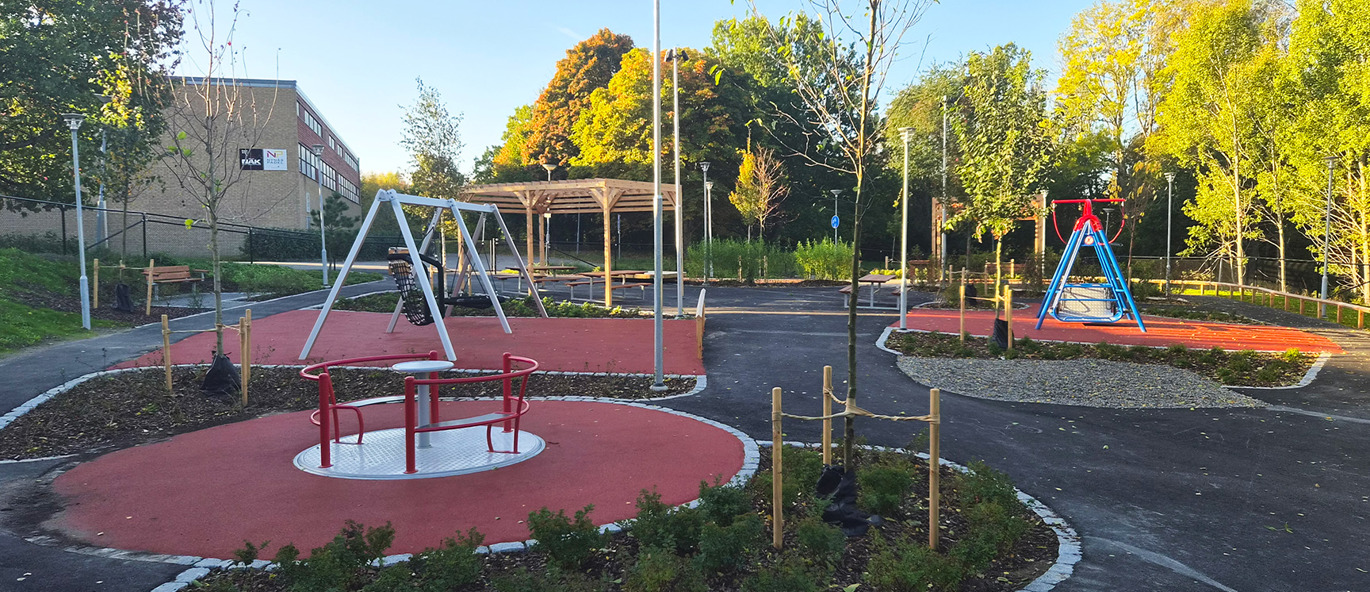 Tillgänglighetsanpassad skolgård på Idun i Nynäshamn med karusell, gungor, sammanhängande gångytor och sittplatser under en pergola, omgiven av planteringar.