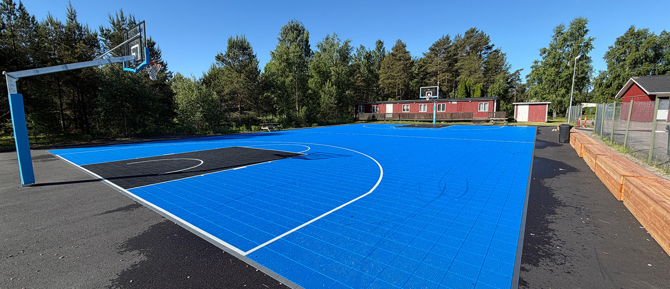 Streetbasketplan på en idrottsplats i Karlstad med blått sportgolv, vita spellinjer, två basketkorgar med genomskinliga plattor och sittmoduler längs ena långsidan.