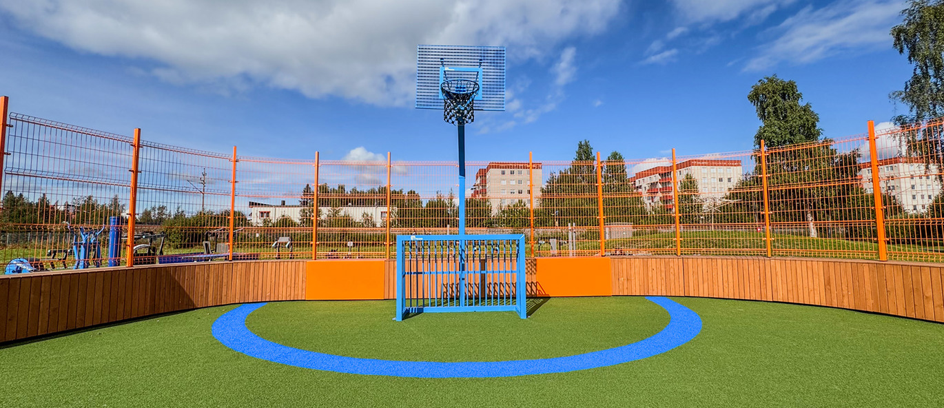 Grön konstgräsmatta med bollsarg och staket av stål och trä i orange färg, bollbål och baskettopp i blått stål.