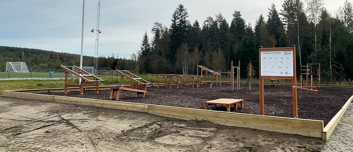 Grusplan och inramat utegym med barkspån på marken, välkomstskylt och träningsstationer av trä.