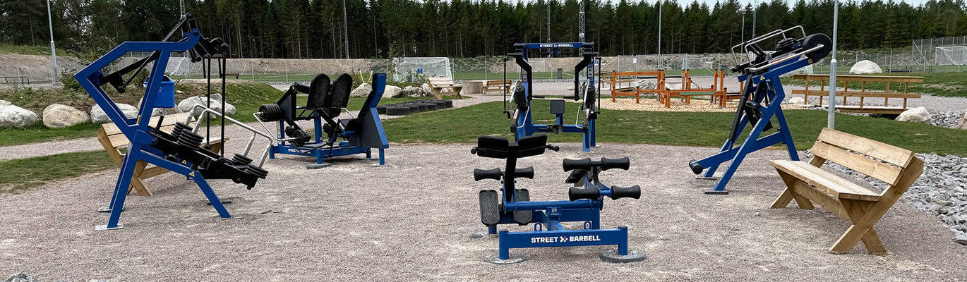 Utegym med styrketräningsutrustning i offentlig miljö intill en idrottsanläggning och grönområde.