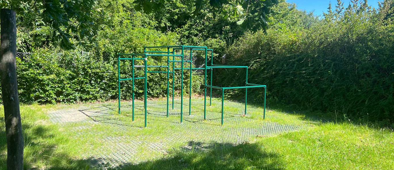 Grønt parkourstativ på et gressområde omgitt av hekker og trær.