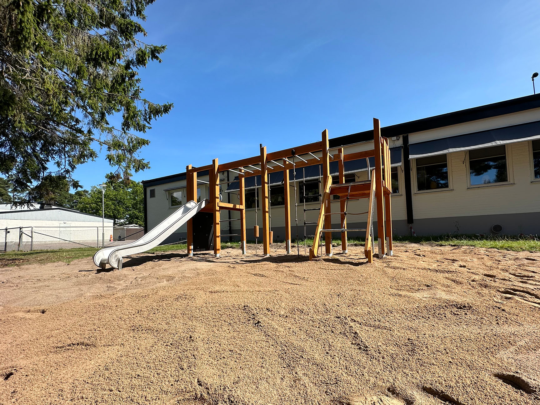 Naturlekplats med träbaserad klätterställning, rutschkana och sandunderlag på en skolgård.