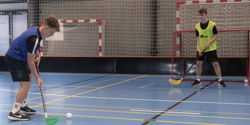 Innebandyspelare med klubba och boll framför ett innebandymål i en idrottshall.