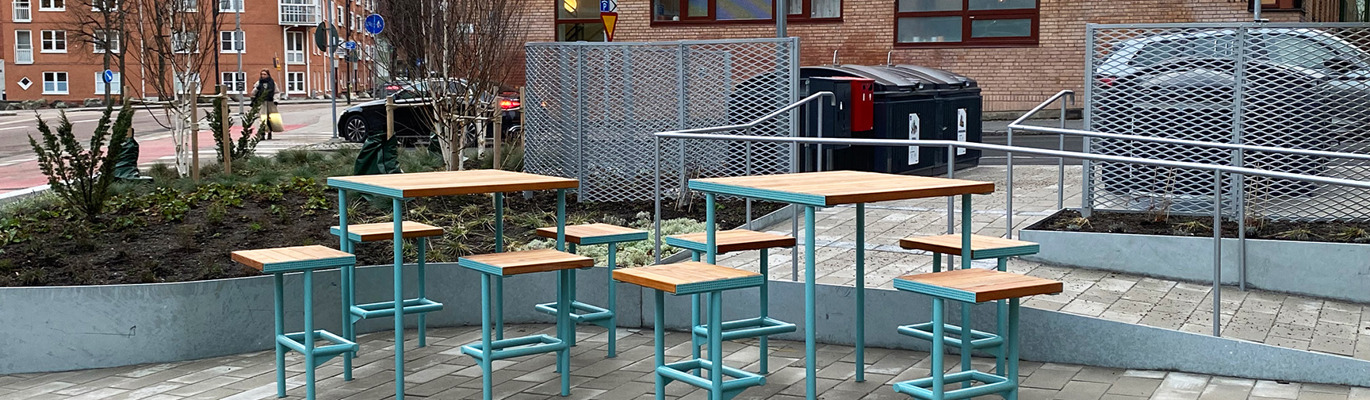 Gestaltad mötesplats med bord, sittplatser och planteringar i offentlig utemiljö som skapar förutsättningar för social samvaro, pauser och vistelse.