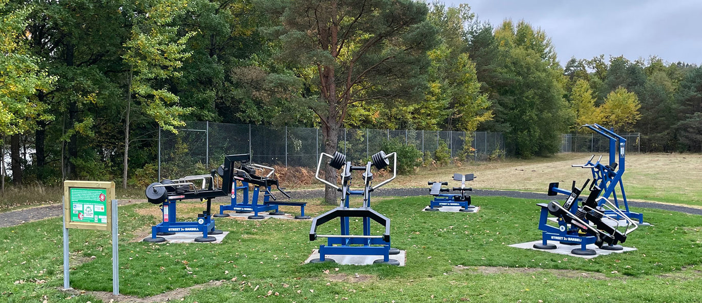 Utegym i Åsljunga med sex blå träningsstationer på en gräsyta, omgivet av träd och gångvägar, med en informationsskylt vid ingången.