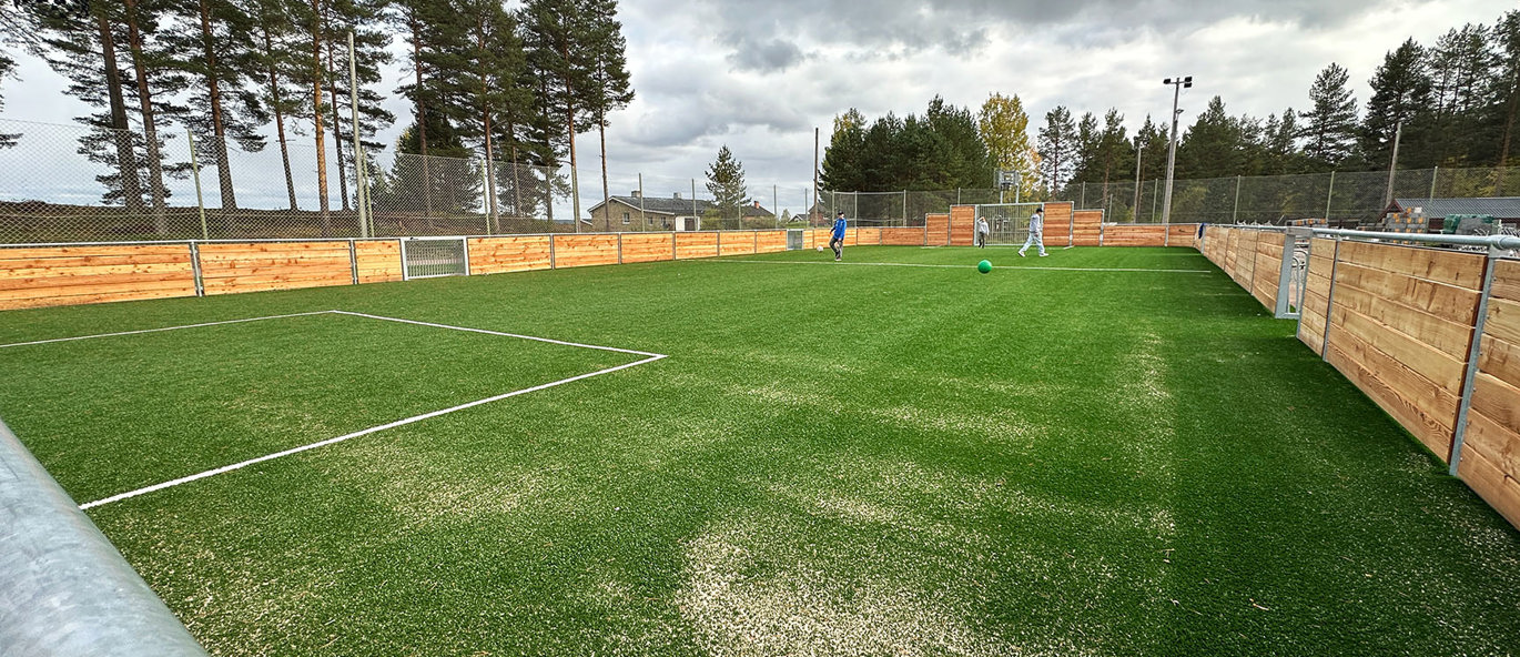 Multiarena med konstgräs på idrottsplats i Mora där barn spelar fotboll, omgiven av träsarg och nätstängsel.