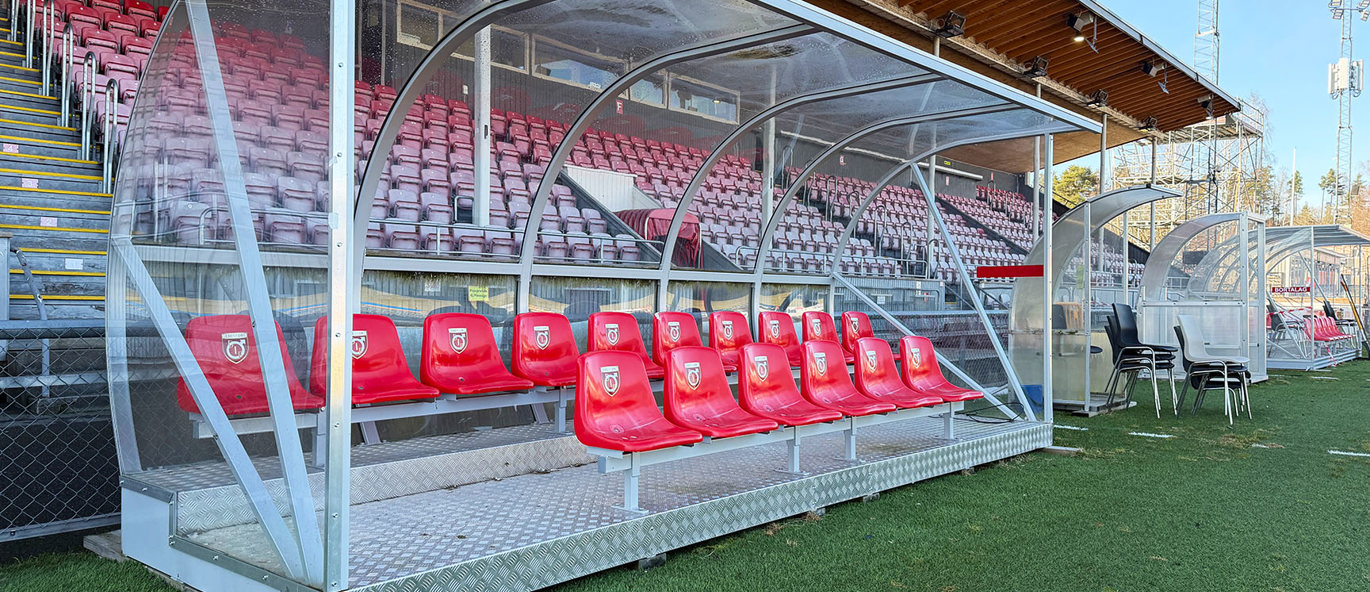 Avbytarbås i transparent polykarbonat med röda sittplatser vid sidlinjen på Stora Valla i Degerfors, med läktare i bakgrunden.