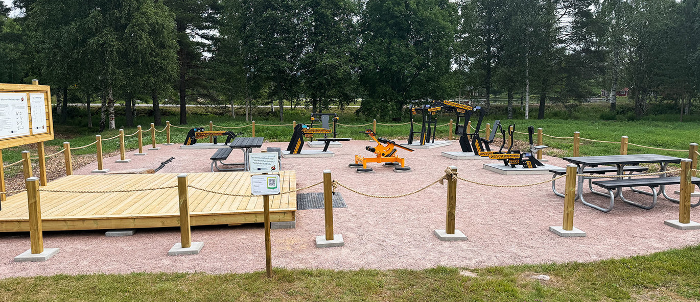 Utegym vid idrottsplatsen i Fredriksberg med svart- och gulfärgade träningsmaskiner, bänkbord och träplattform omgivet av grönska.