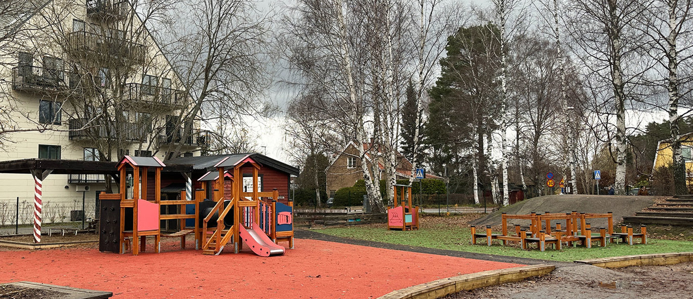 Lekplats med rött gummiunderlag och grön gräsmatta, klätterställning, lekhus och balansbana.