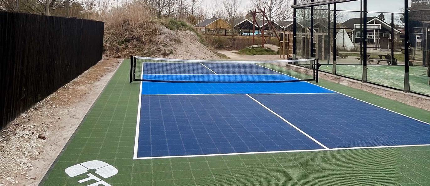 Blå pickleballbane med blått nett på grønt underlag, med Tress-logoen i hvitt, og en padelbane i bakgrunnen.