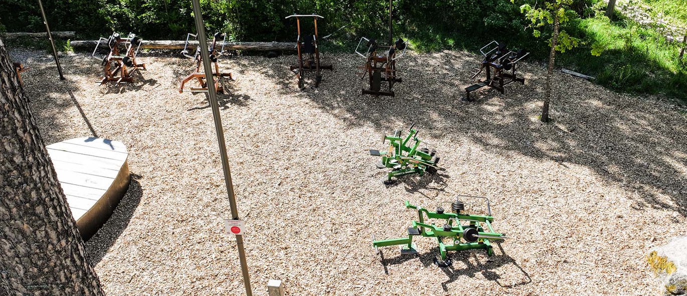 Flygfoto över utegymmet i Vivalla, Örebro, med Street Barbell-träningsmaskiner i gröna och bruna kulörer placerade i en naturnära parkmiljö omgiven av tallar och träflisunderlag.