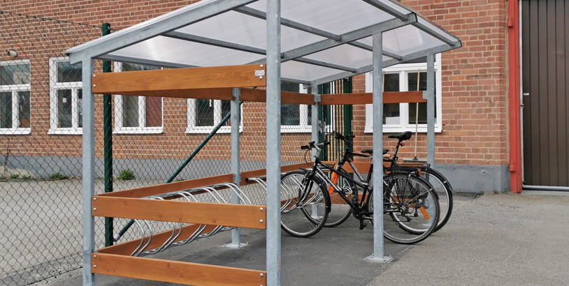 Cykelgarage med transparent tak, stålkonstruktion och trädetaljer som skyddar parkerade cyklar mot väder och vind.