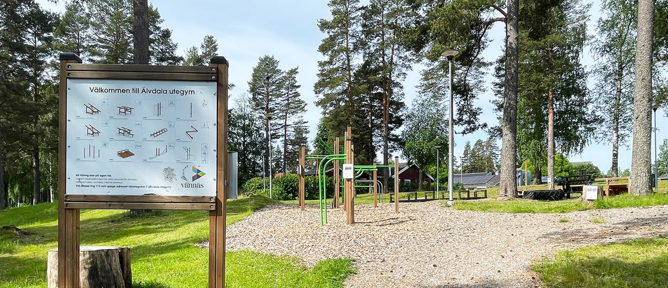 Informationsskylt med bilder på träningsstationer i en park med ett utegym med olika redskap.