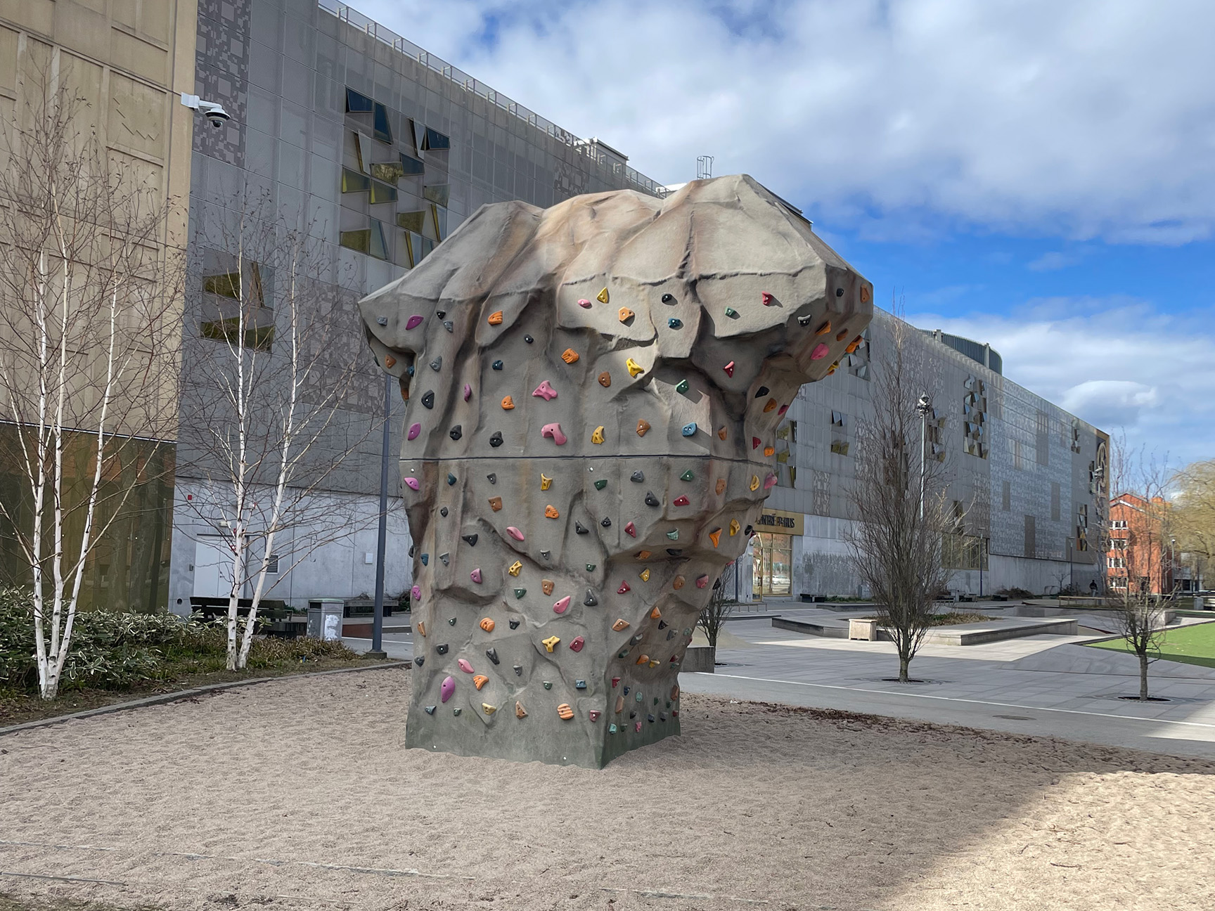 Klätterstenen Boulder Rocks Bergstoppen på lekplats vid galleria i Kristianstad, en stor skulptural klättersten med färgglada klättergrepp placerad i stadsmiljö.