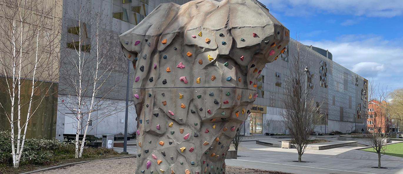 Buldresteinen Proff på en lekeplass ved et kjøpesenter i Kristianstad – en stor, skulpturell klatrestein med fargerike klatregrep plassert i et urbant miljø.