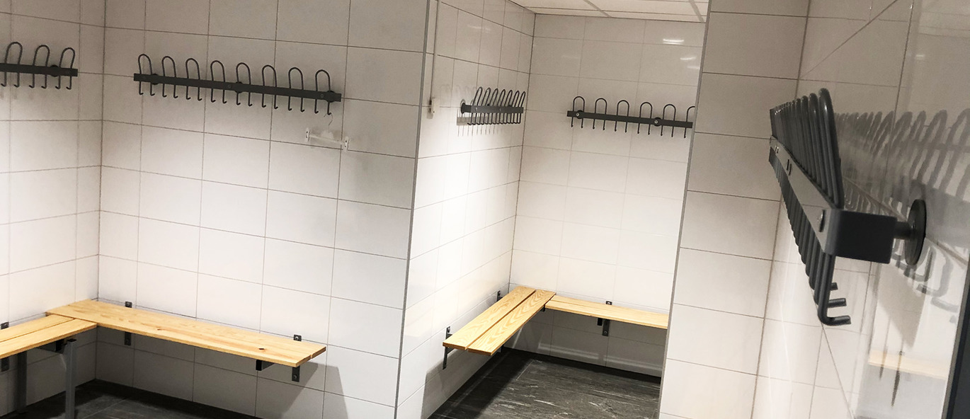 Omklädningsrum i padelhall med väggmonterade svarta krokar och träbänkar längs kaklade väggar i en ljus och avskalad miljö.