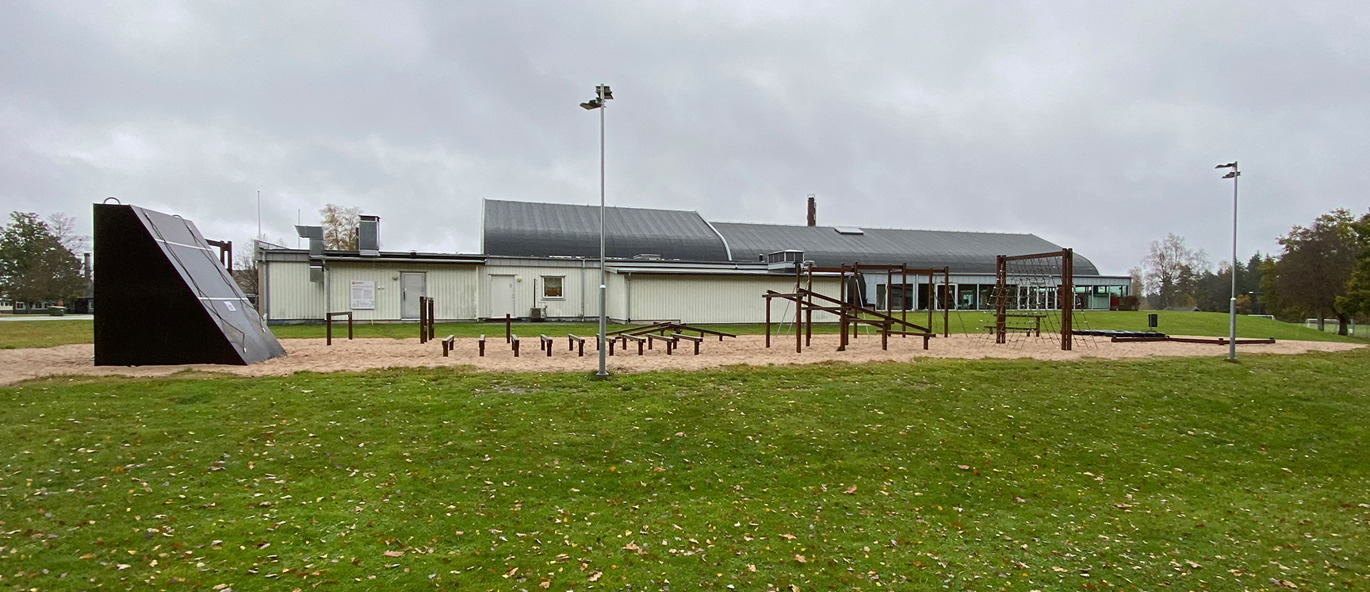 Hinderløype på grus med mange hinder i tre. Gressplen og idrettshall i bakgrunnen.