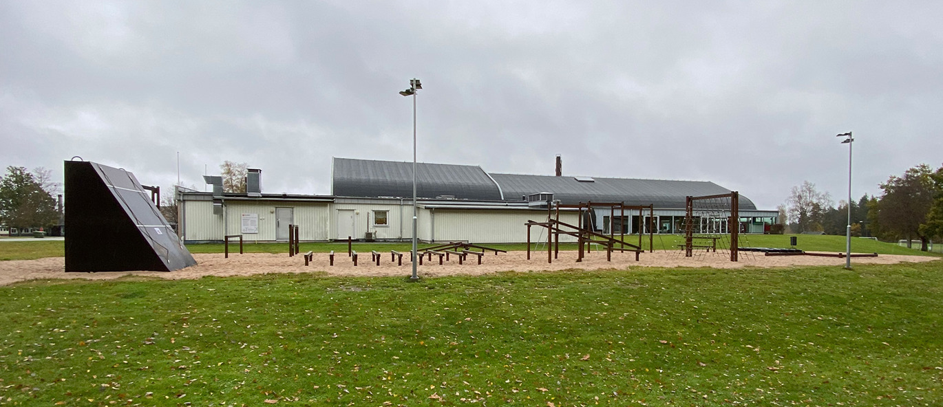 Hinderbana på grus med många hinder i trä. Gräsmatta och sporthall i bakgrunden.