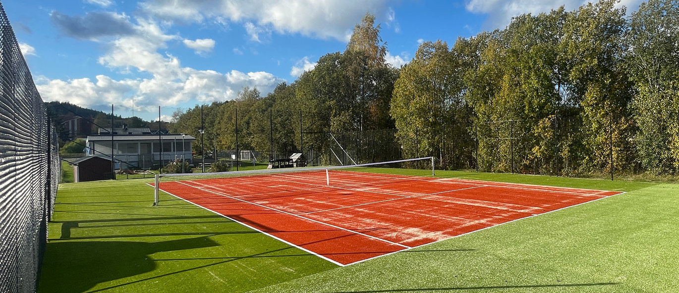 Grönt konstgräs, röd tennisplan med vita linjer, skog i bakgrunden och staket runt om.