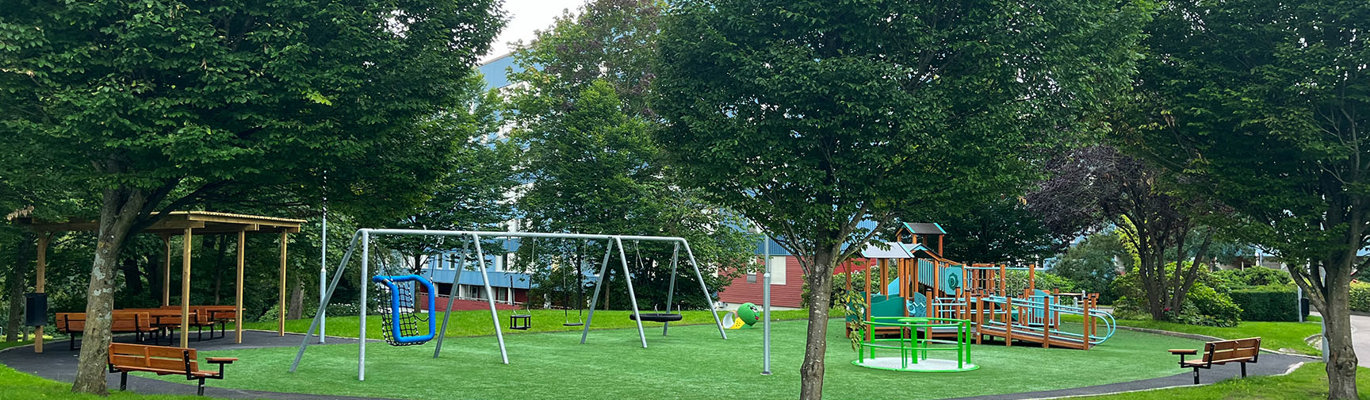 Tillgänglighetsanpassad lekplats i grön parkmiljö med gungor, breda gångar, marknära lekutrustning och sittplatser under träd och pergola.