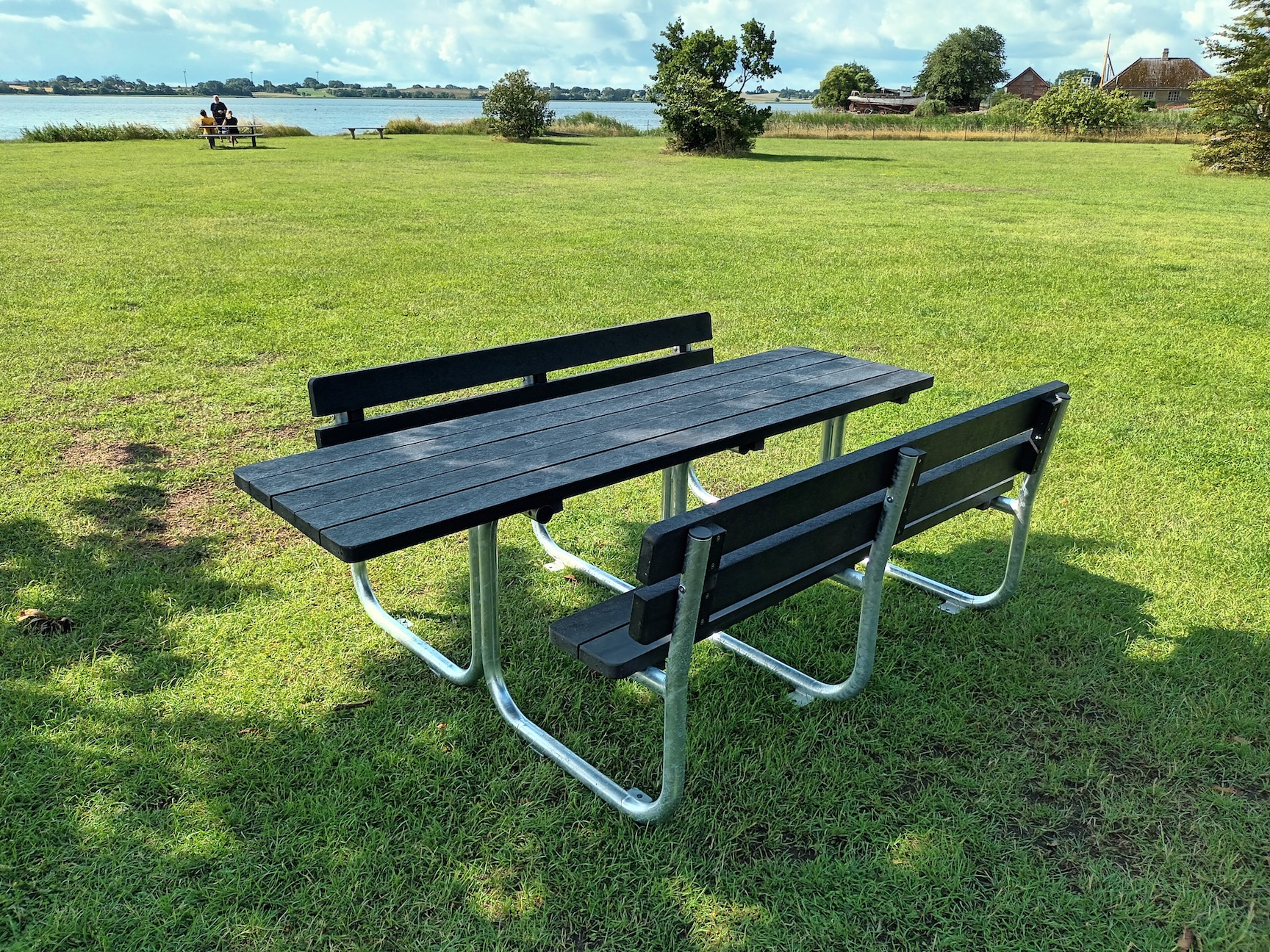 Tillgänglighetsanpassat parkbord med sittbänkar i svart trä och stabilt metallstativ placerat på gräsyta.