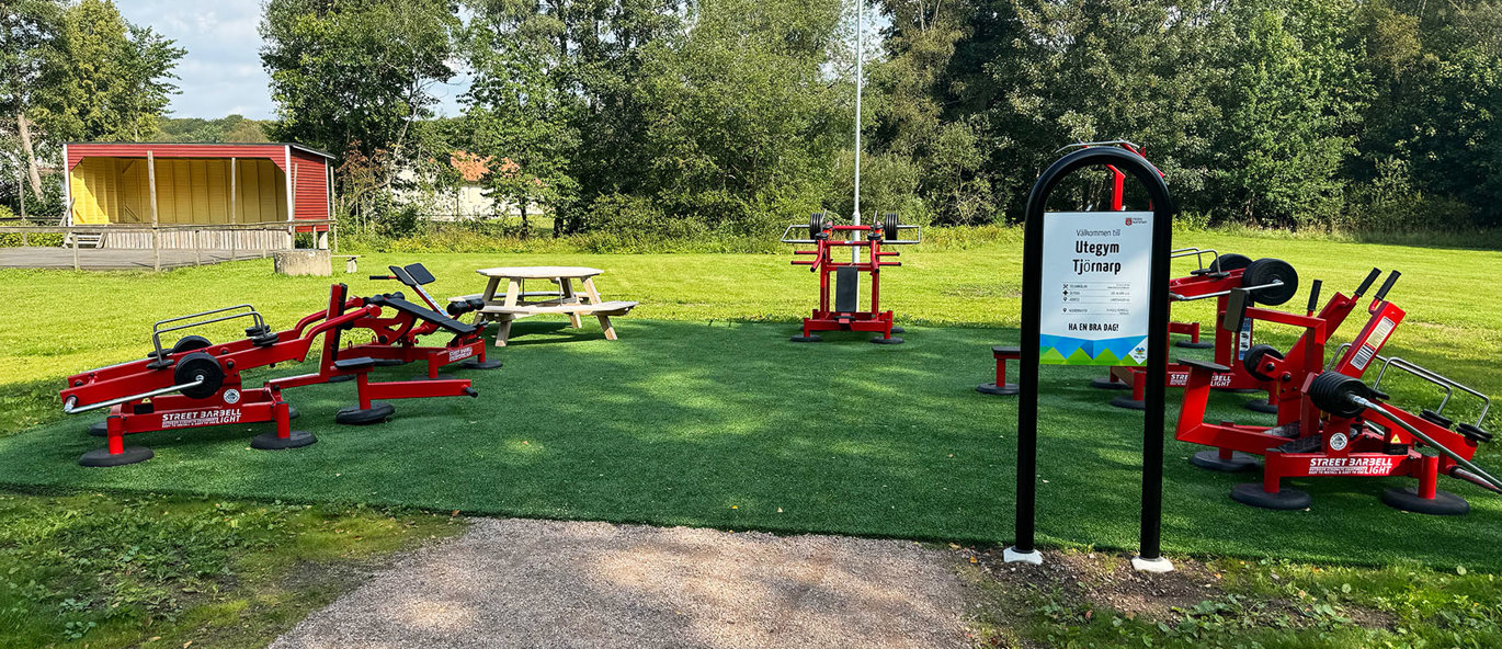 Grön gräsmatta i en park med röda träningsmaskiner och en välkomstskylt.