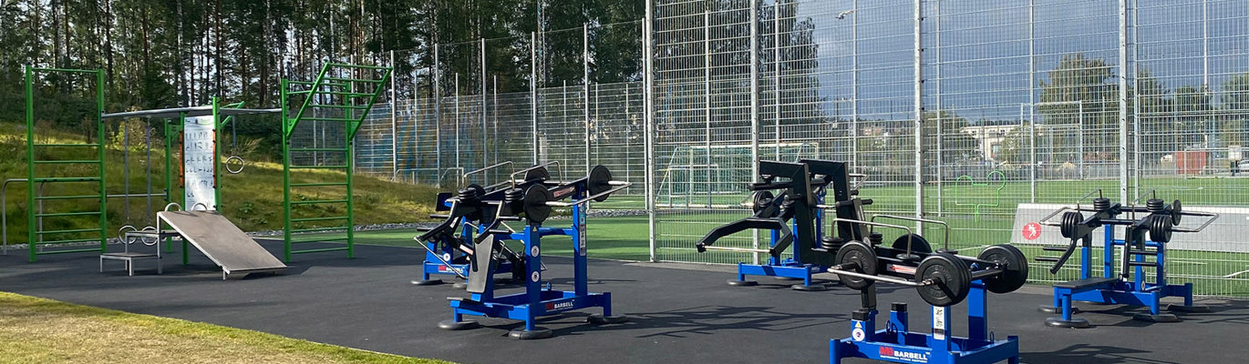 Utomhusgym och träningsyta vid idrottsanläggning med styrketräningsutrustning, hinderbana och inhägnad konstgräsplan.