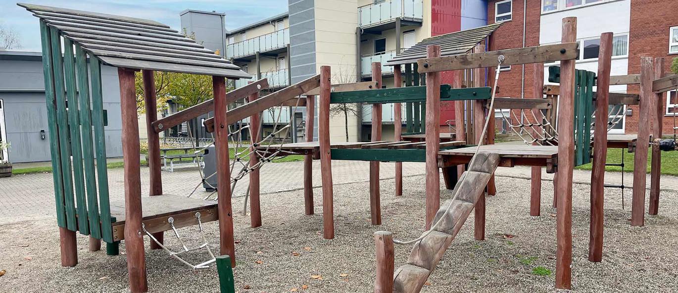 Legeplads med klatreleg på grusbelægning med boliger i baggrunden