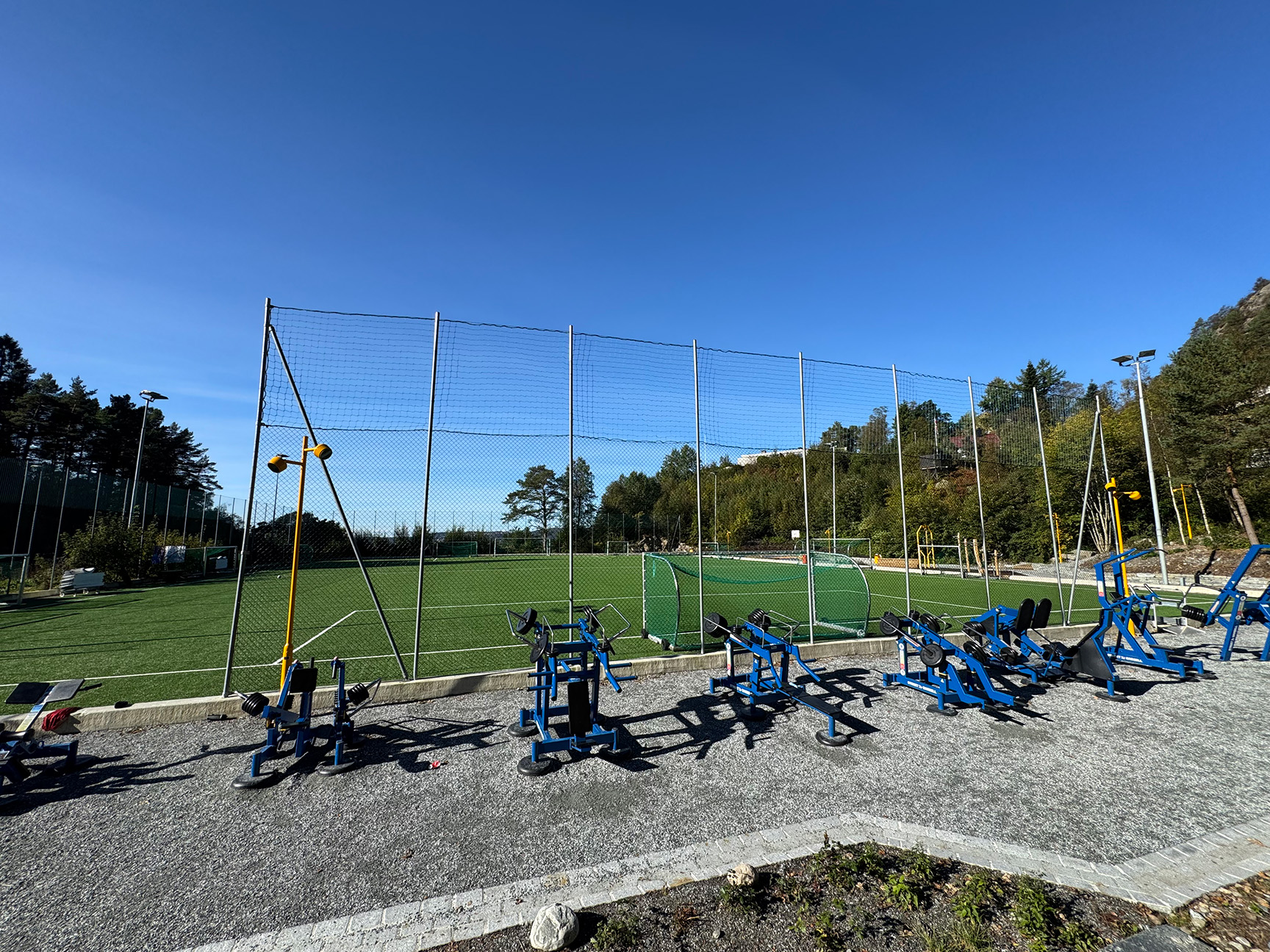 Blåa träningsmaskiner på en grusplan vid en fotbollsplan.