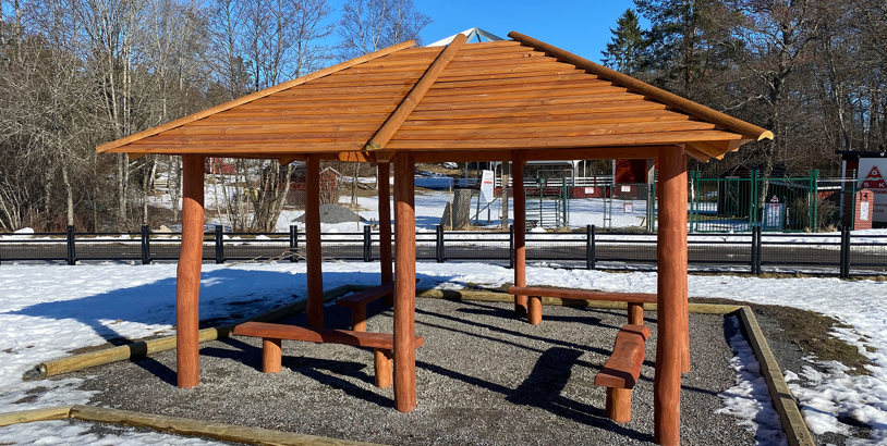 Öppet grillhus i trä med tak och fasta bänkar, placerat på grusyta i parkmiljö under vintertid.