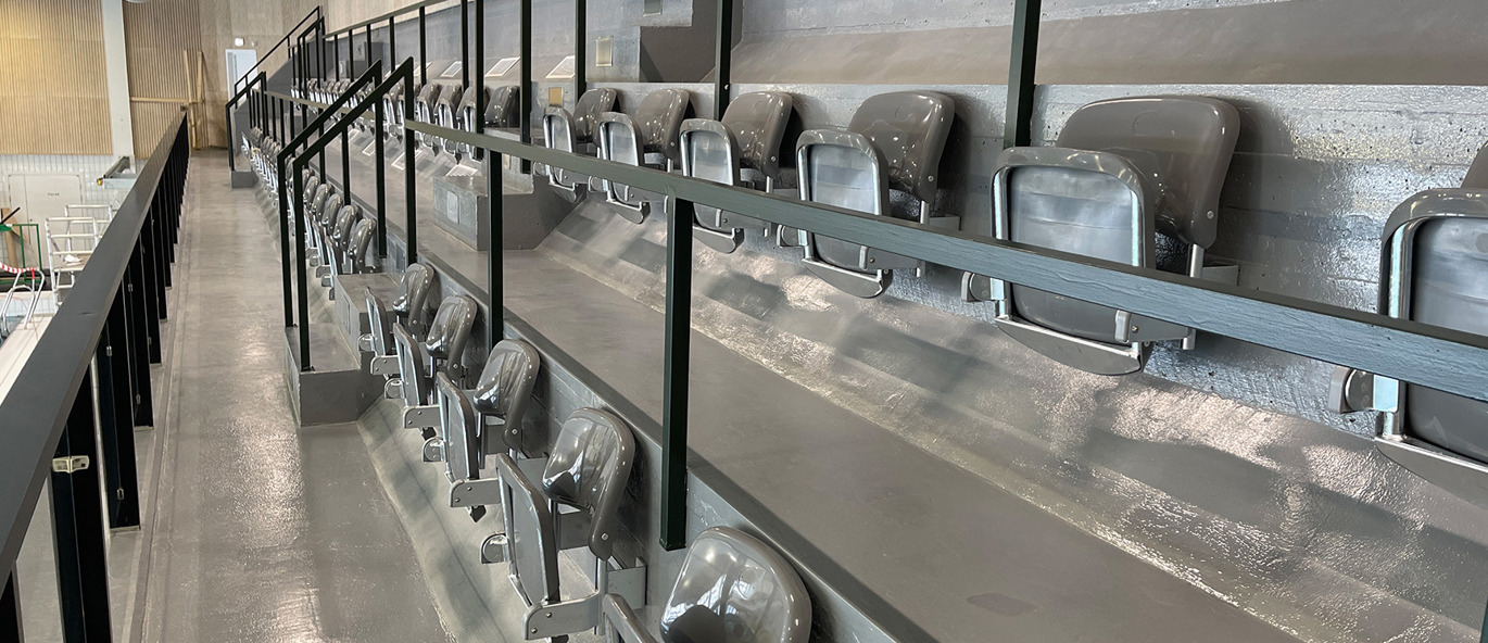 Läktarstolar i ljusgrått monterade på betongläktare i modern simhall i Älvsbyn, med fällbara sitsar och räcken längs gångstråk