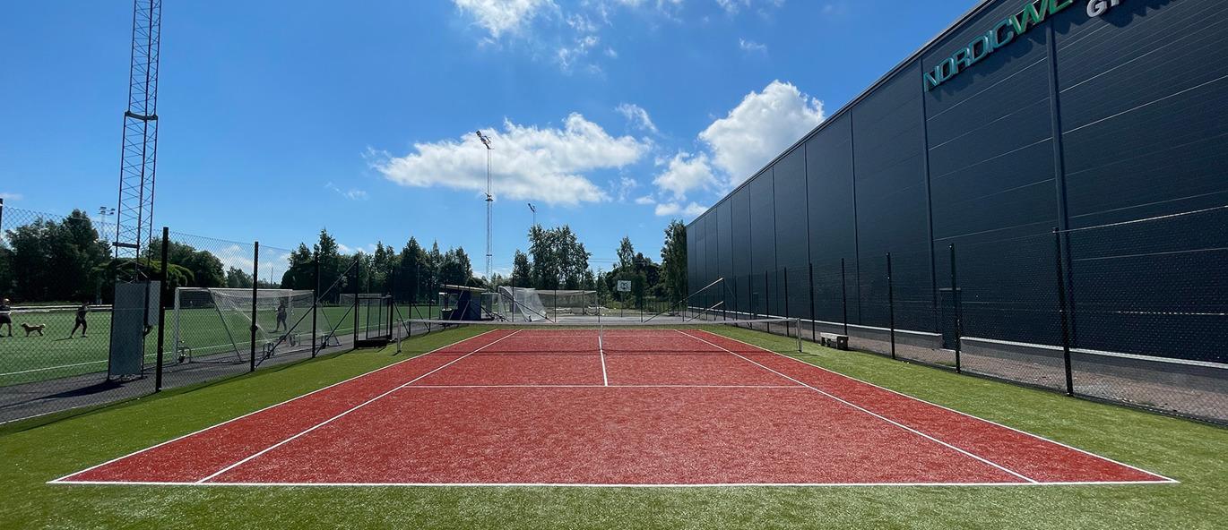 Tennisplan med konstgräs i färgerna rött, grön och vitt framför en idrottshall.