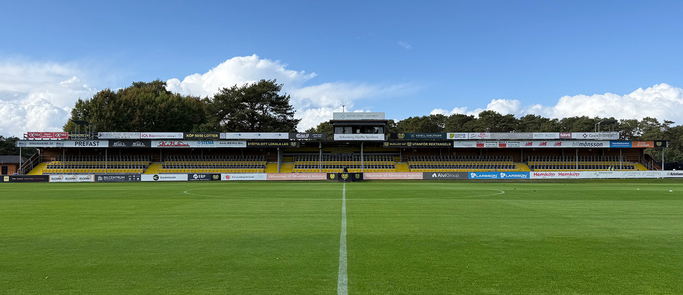 Strandvallen i Hällevik, Mjällby AIF:s hemmaarena, med fotbollsplan och taktäckt läktare med gula och svarta Safe Standing-läktarstolar.