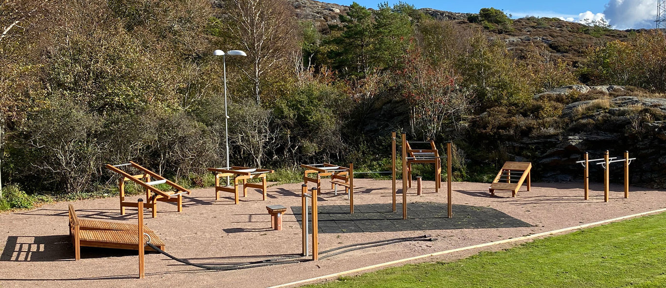 Grön fotbollsplan framför ett utegym med 10 olika träningsstationer av trä, träd och grönska i bakgrunden.