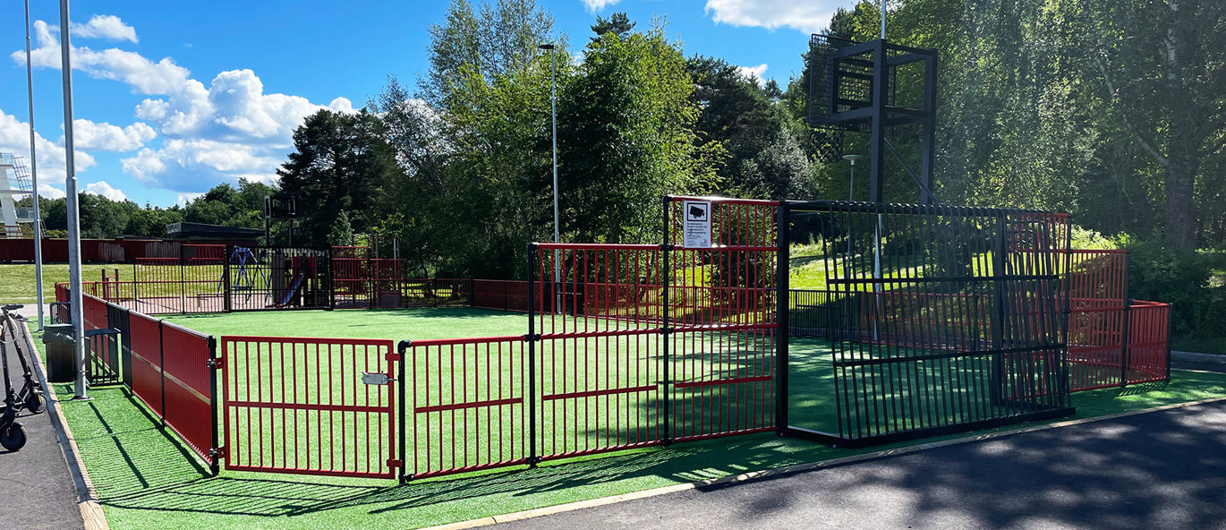 Multibane i rød og sort med grønt kunstgræs, kantafskærmning omkring og store mål og basketmål på de korte sider