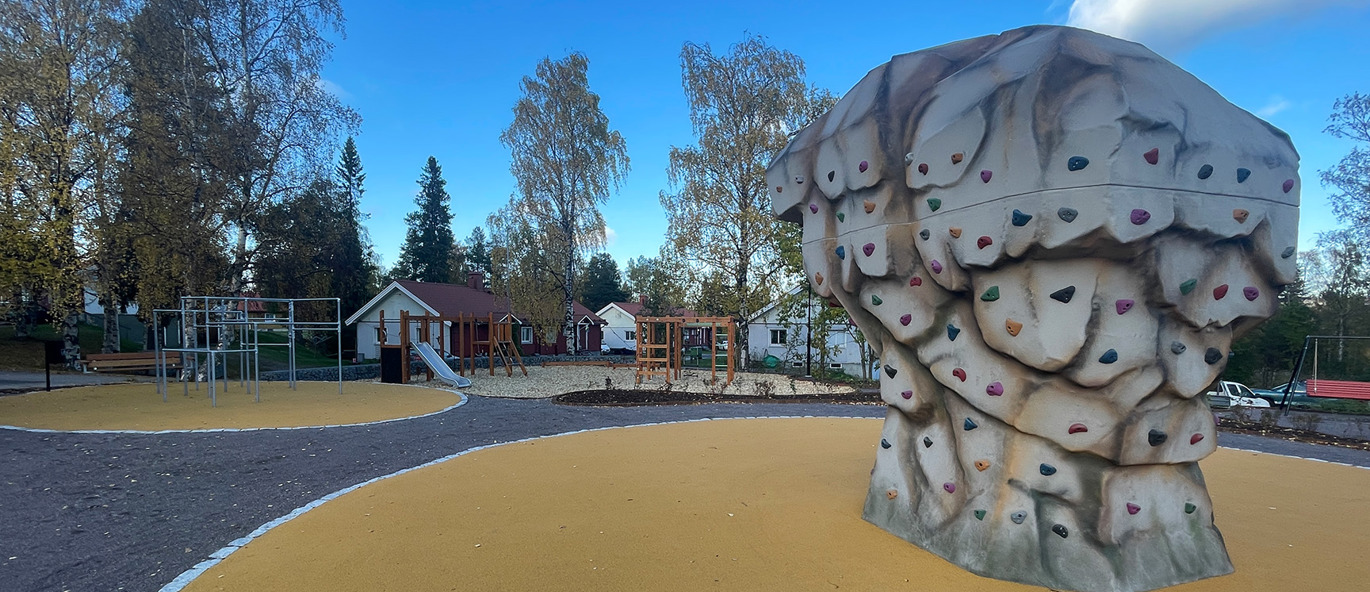 Stor klättersten på gult gummiunderlag, med en lekplats i bakgrunden.