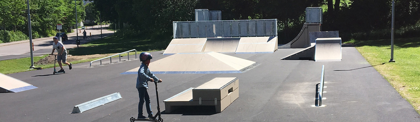 Skatepark med ramper, rails och åkytor där barn och ungdomar använder kickbike i en aktiv utemiljö.
