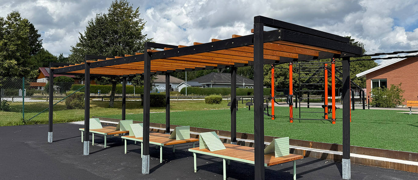 Stor pergola med svarte stolper og tak av trespiler, plassert på asfalt sammen med tre loungebenker i tre og grønnlakkert stål.
