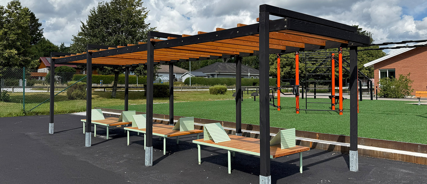 Stor pergola med svarta stolpar och träribbor som tak, står på asfalt tillsammans med tre loungebänkar av trä och grön metall.