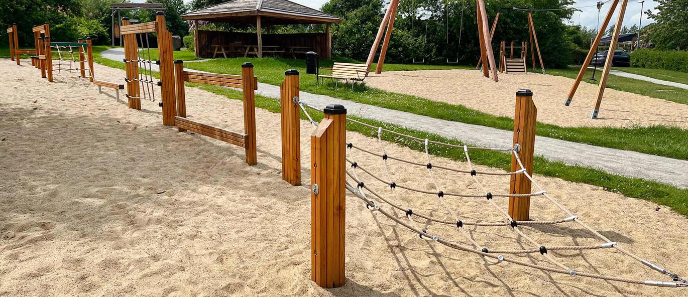 Lekeplass med grusdekke og lekeapparater i tre, inkludert balansebaner, huskestativer, bålhus og klatrenett.
