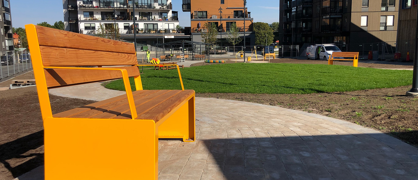 En gul parksoffa på en stenlagd gång med bostadshus i bakgrunden.