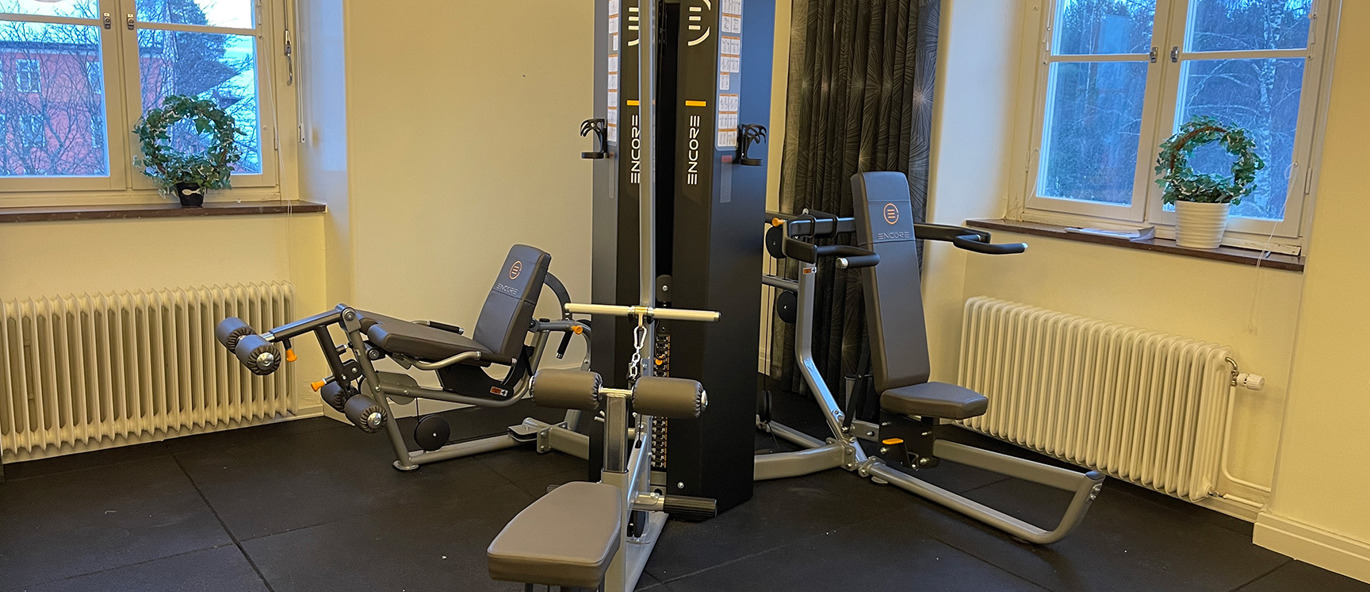 Multigym med träningsbänk i ljust gymrum med gummigolv och fönster.