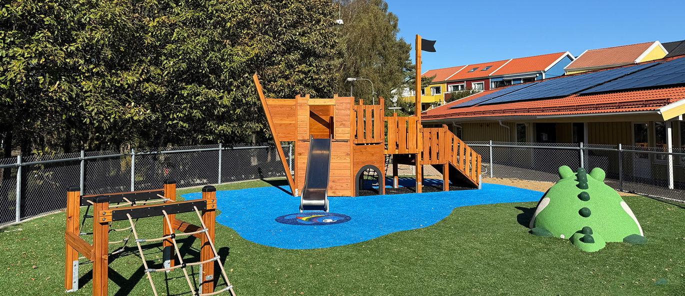 Grønn gressplen med et klatrestativ i form av en båt, et klatrenett og en gummifigur som forestiller en krokodille.